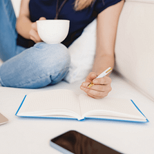 woman writing on notebook with blue cover, holding coffee on white couch