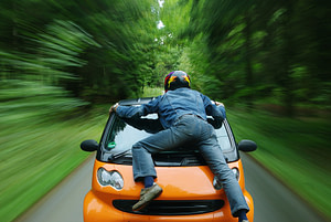 Man clinging to a car hood while it speeds down a forest highway